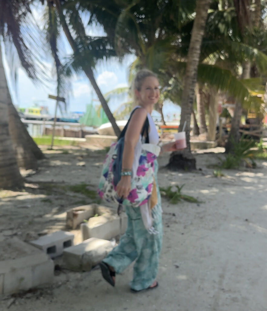 blonde-woman-with-bright-beach-bag-and-turquoise-pants-carrying-tropical-juice-in-front-of-palm-trees-in-belize