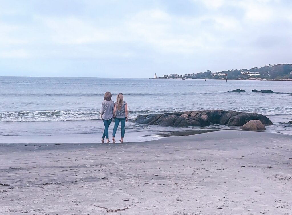 mom-and-daughter-in-jeans-and-gray-shirts-standing-at-waters-edge-on-boston-north-shore-Wingaersheek-Beach