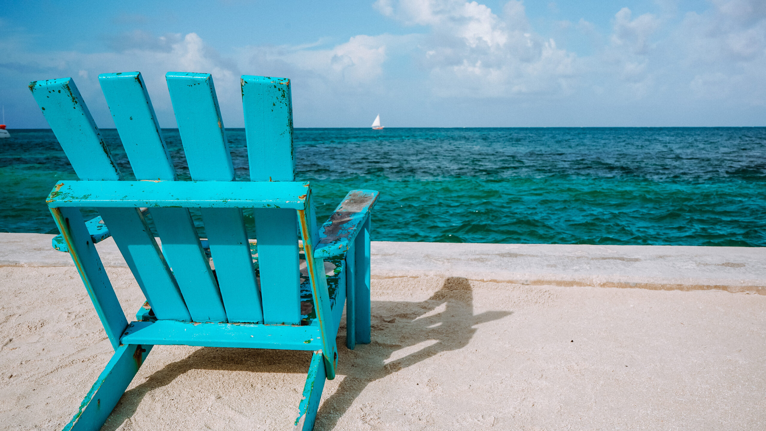 bright-turquoise-adirondack-chair-overlooking-deep-azure-turquoise-sea-with-sailboat-in-the-background