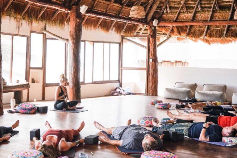 A circle of yoga students lying on their mats with an instructor sitting down, her hand on her heart.