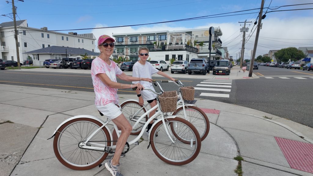A woman in a pink hat and shirt and a teenage boy in a white shirt on white cruiser bikes in front of a hotel in Wildwood NJ