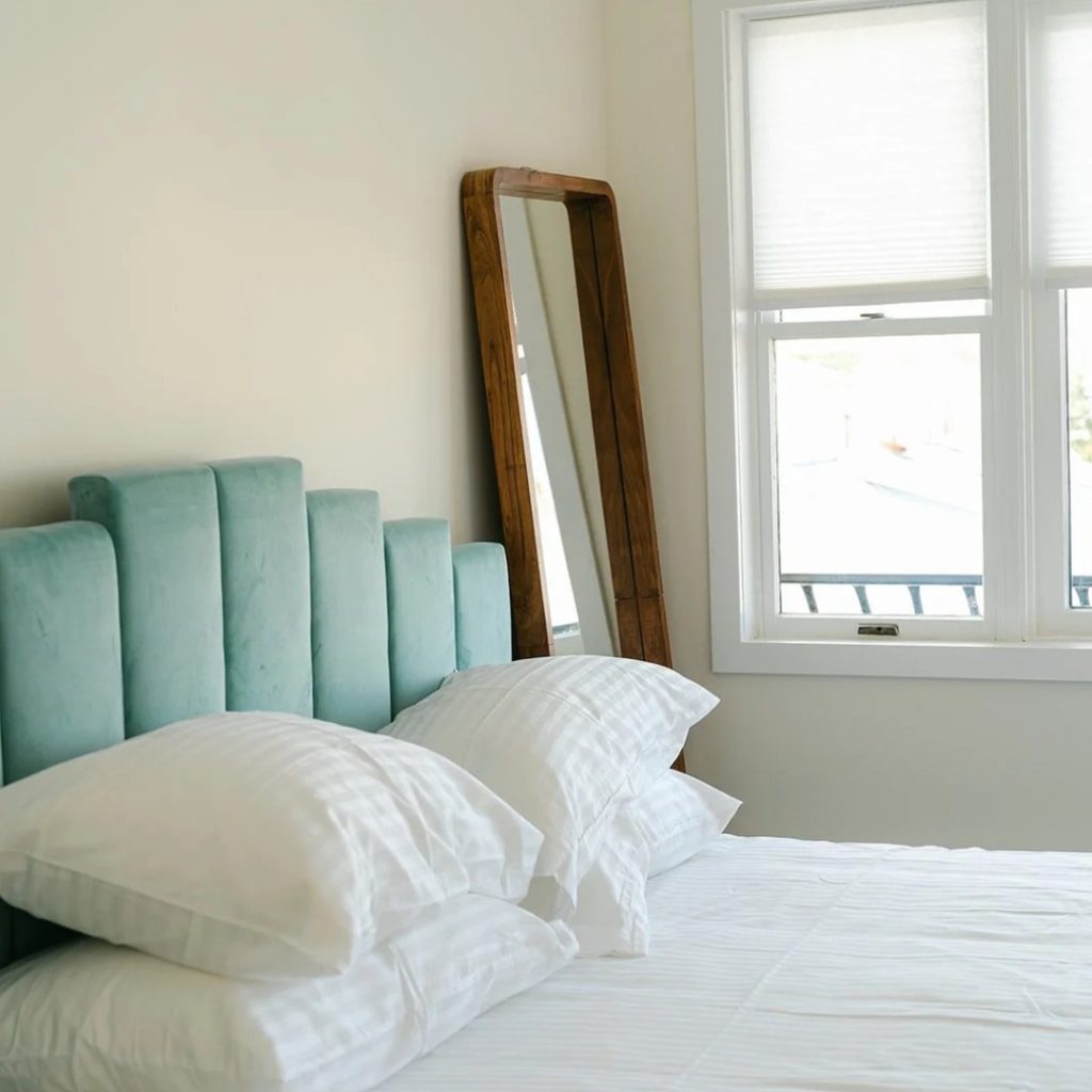 A hotel bed with a turquoise frame and a wooden-framed mirror next to it, with an open window next to that.