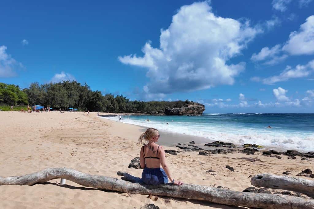 A woman in blue shorts and a bathing suit sitting on driftwood on a beach with some swimmers on it and trees in the background.