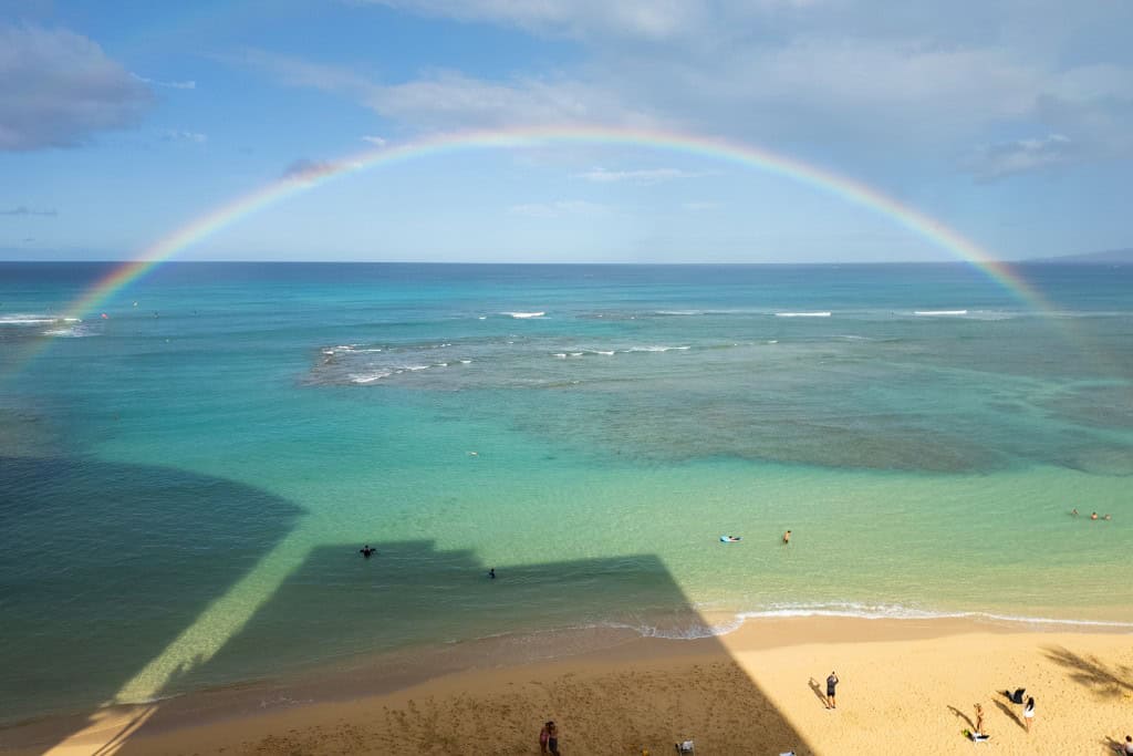 A full rainbow over calm turquoise water and a few people in the water