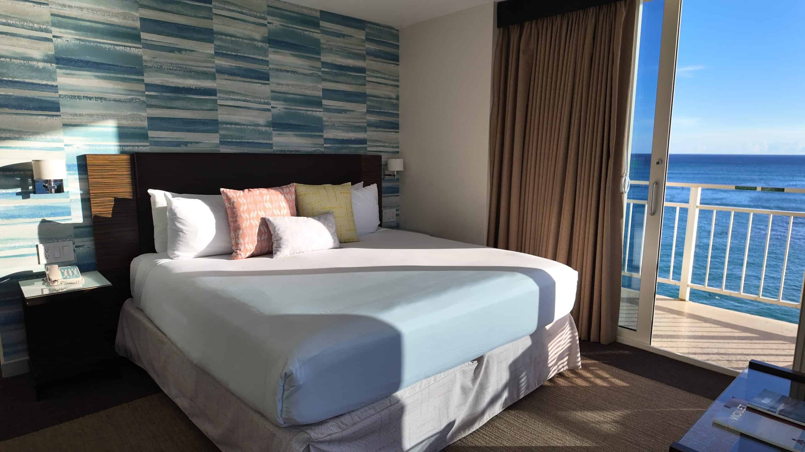 A hotel bed with sunlight on it and an ocean view patio.
