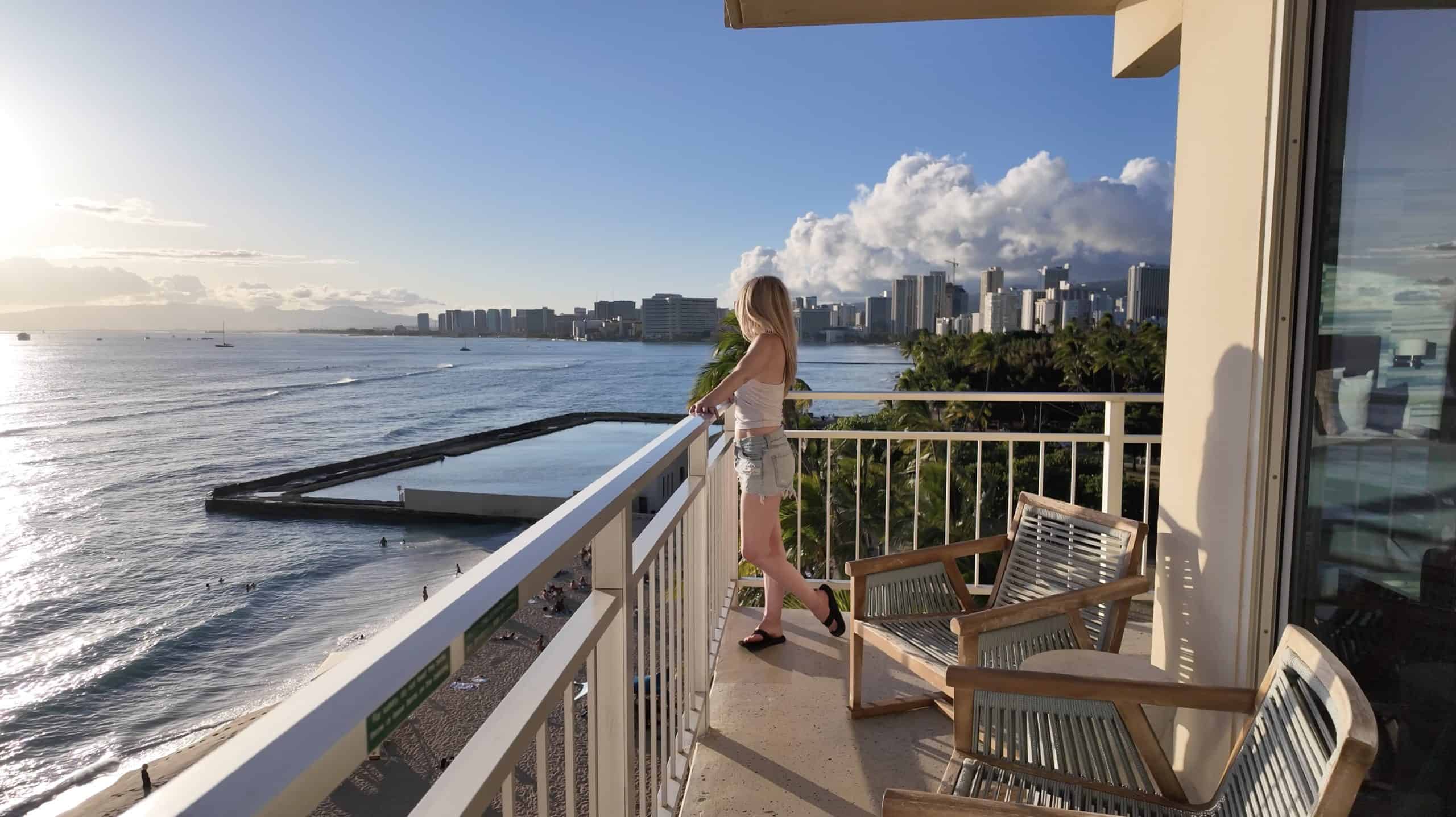 A blonde woman overlooking Waikiki at sunset from a Kaimana Beach Hotel oceanfront balcony with panoramic views of Honolulu.