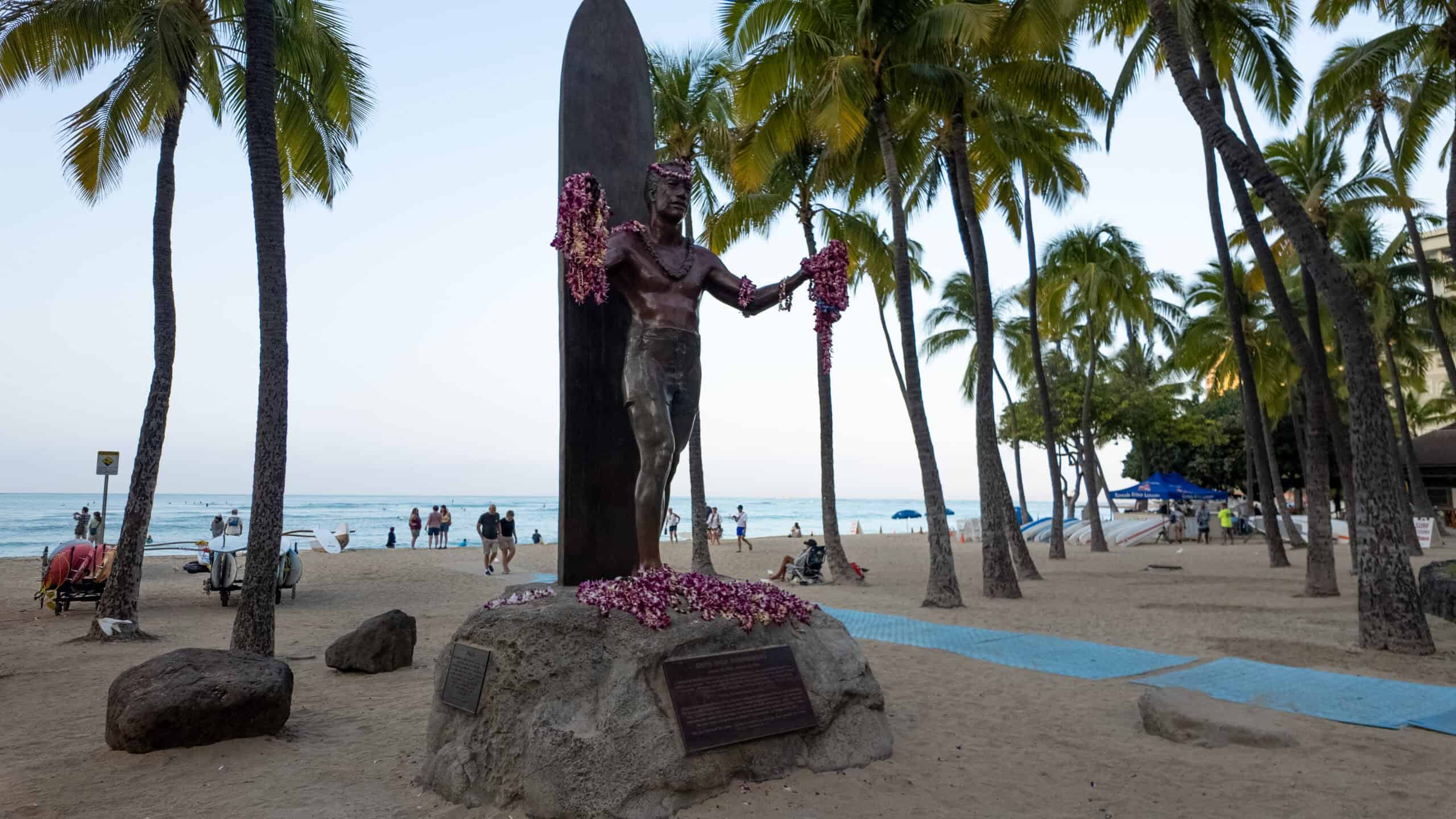 The iconic statue of Duke Kahanamoku holding gifted leis with Waikiki Beach behind it.