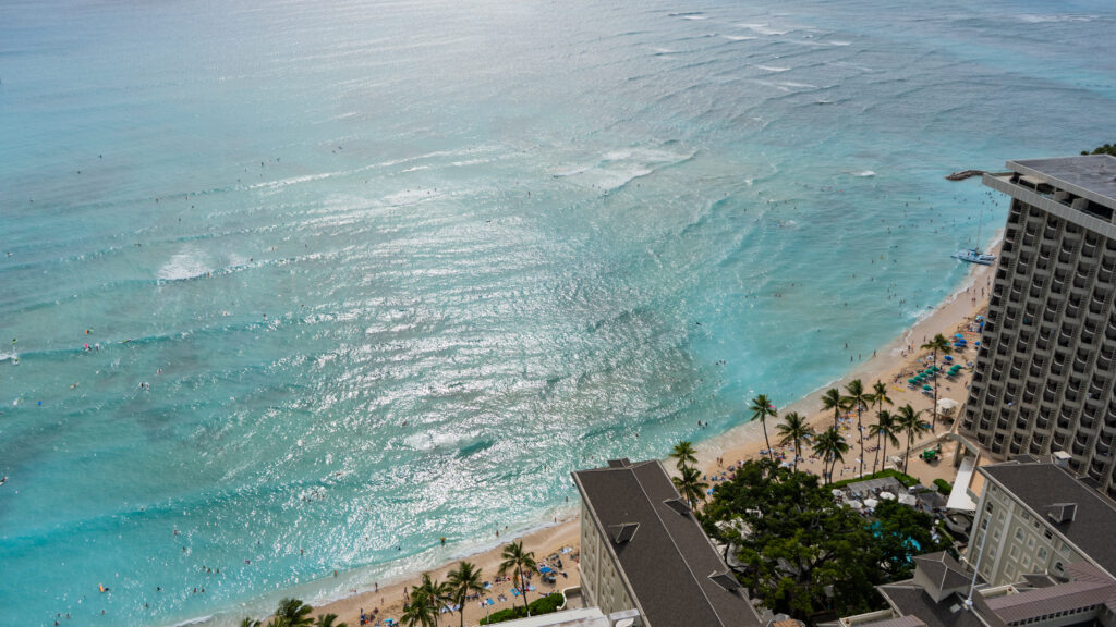 An aerial view over Waikiki Beach showing a turquoise ocean, surfers and palm trees on the sand