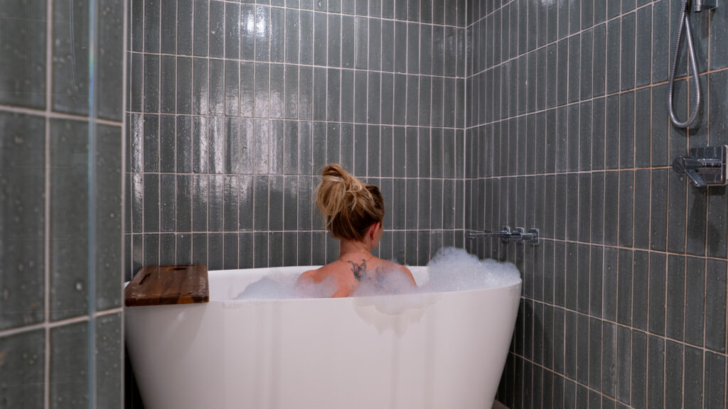 A blonde woman in a white soaking tub full of bubbles in a blue tiled shower