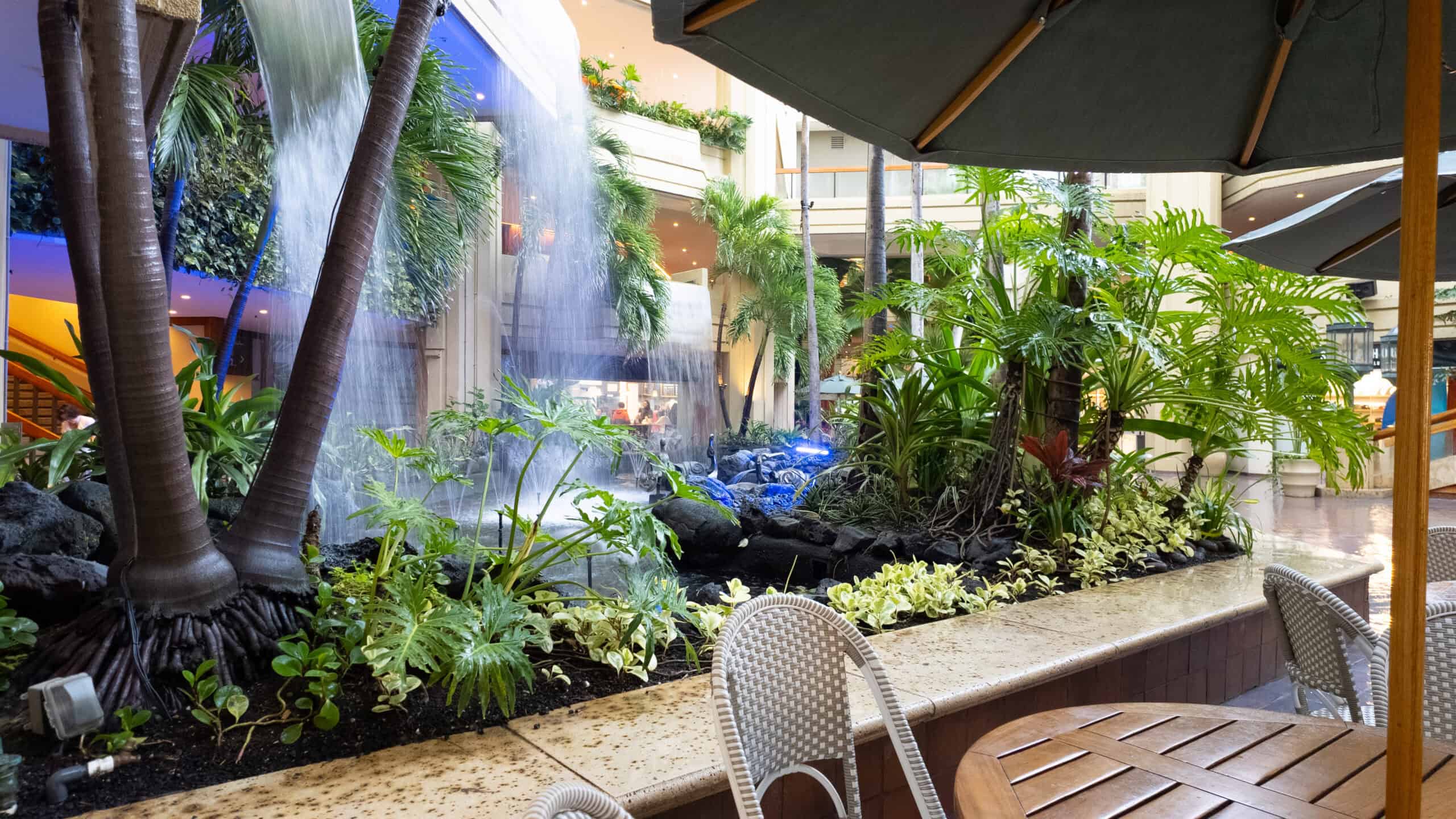 A waterfall surrounded by tropical plants with a table, umbrella and chairs in the forefront