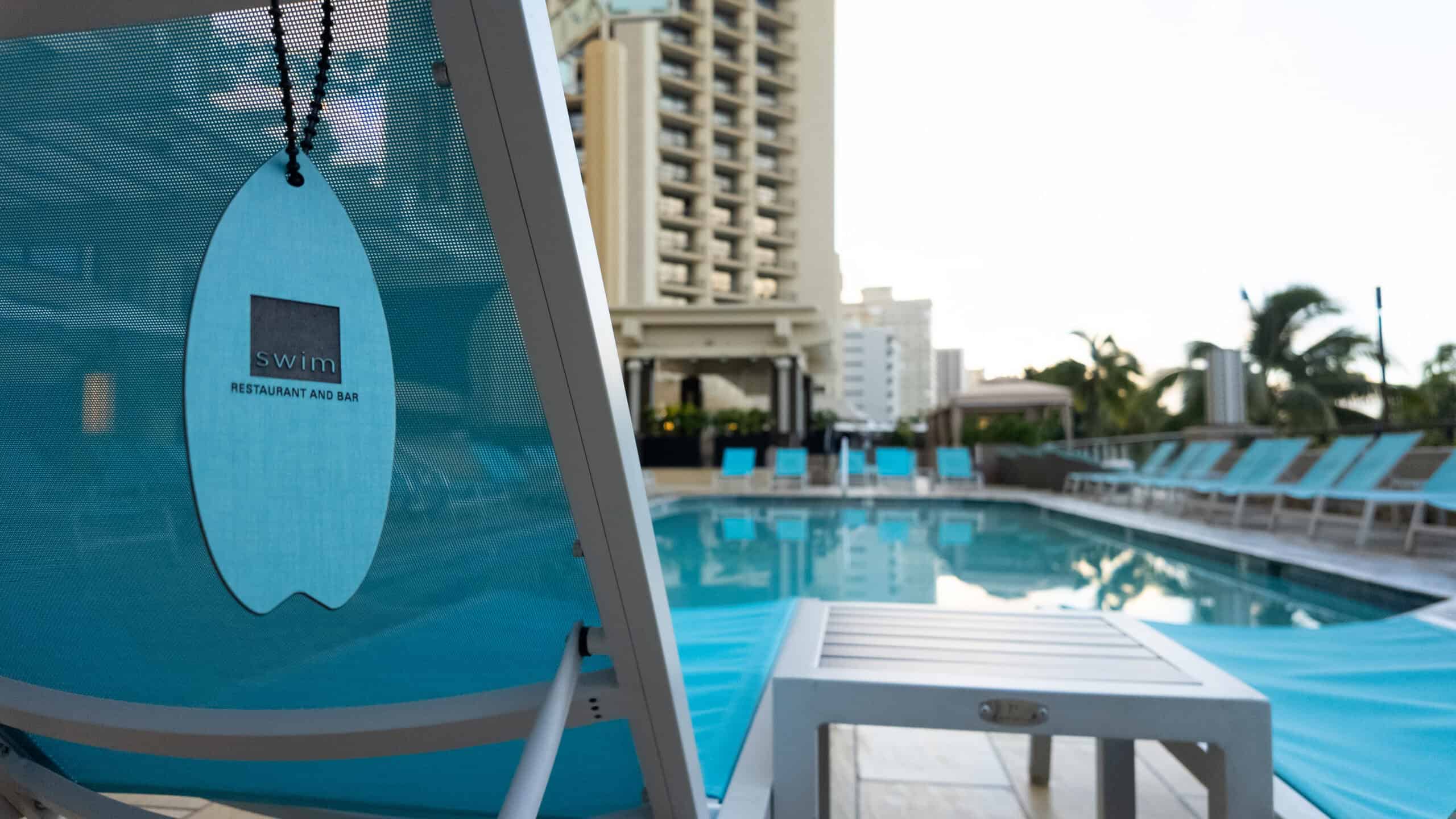 A blue chaise lounge chair with a tag that says SWIM Restaurant on it, and a pool in the background