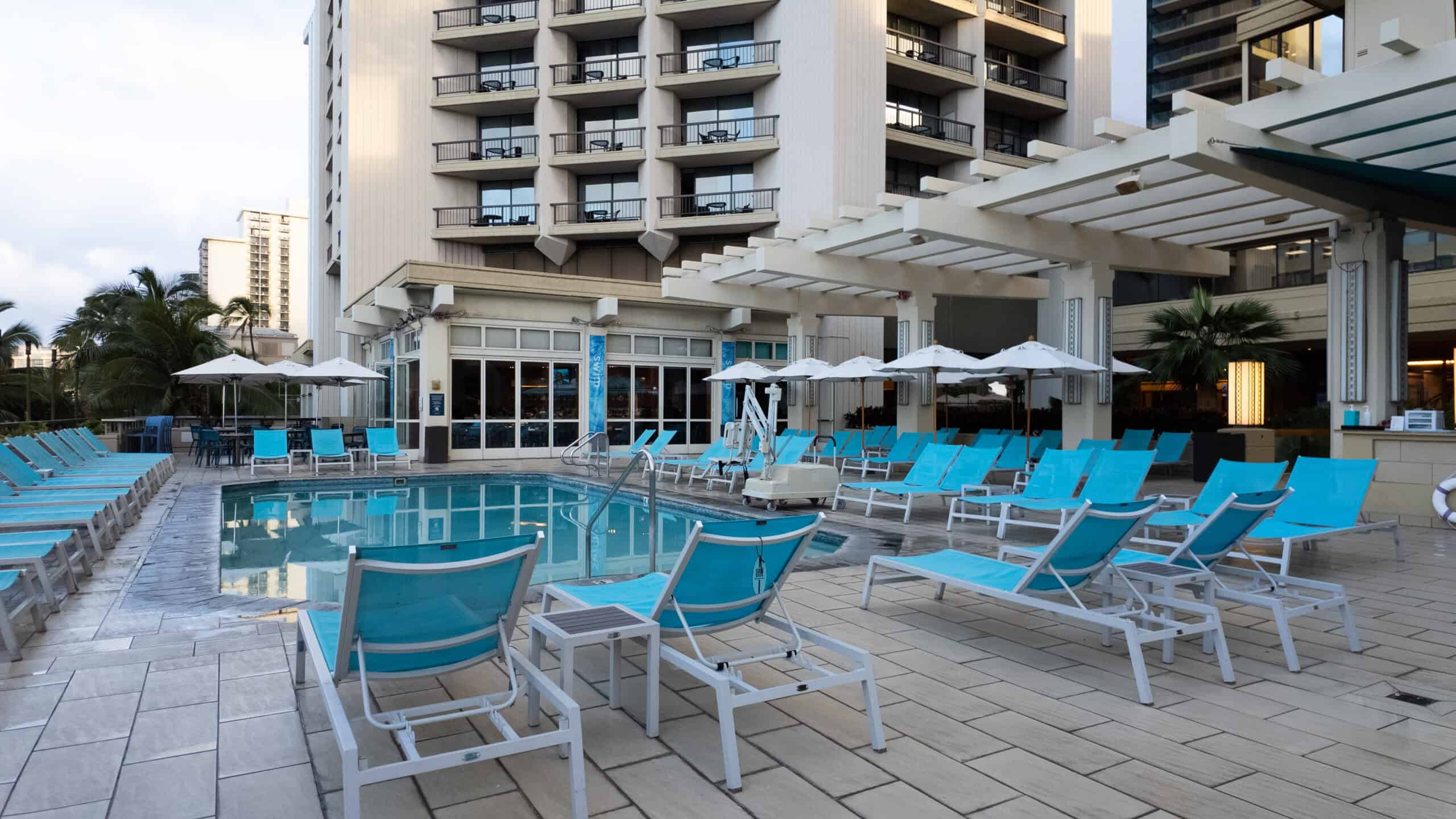 A hotel pool surrounded by turquoise chaise lounge chairs and white umbrellas with a sign for SWIM Restaurant in the background