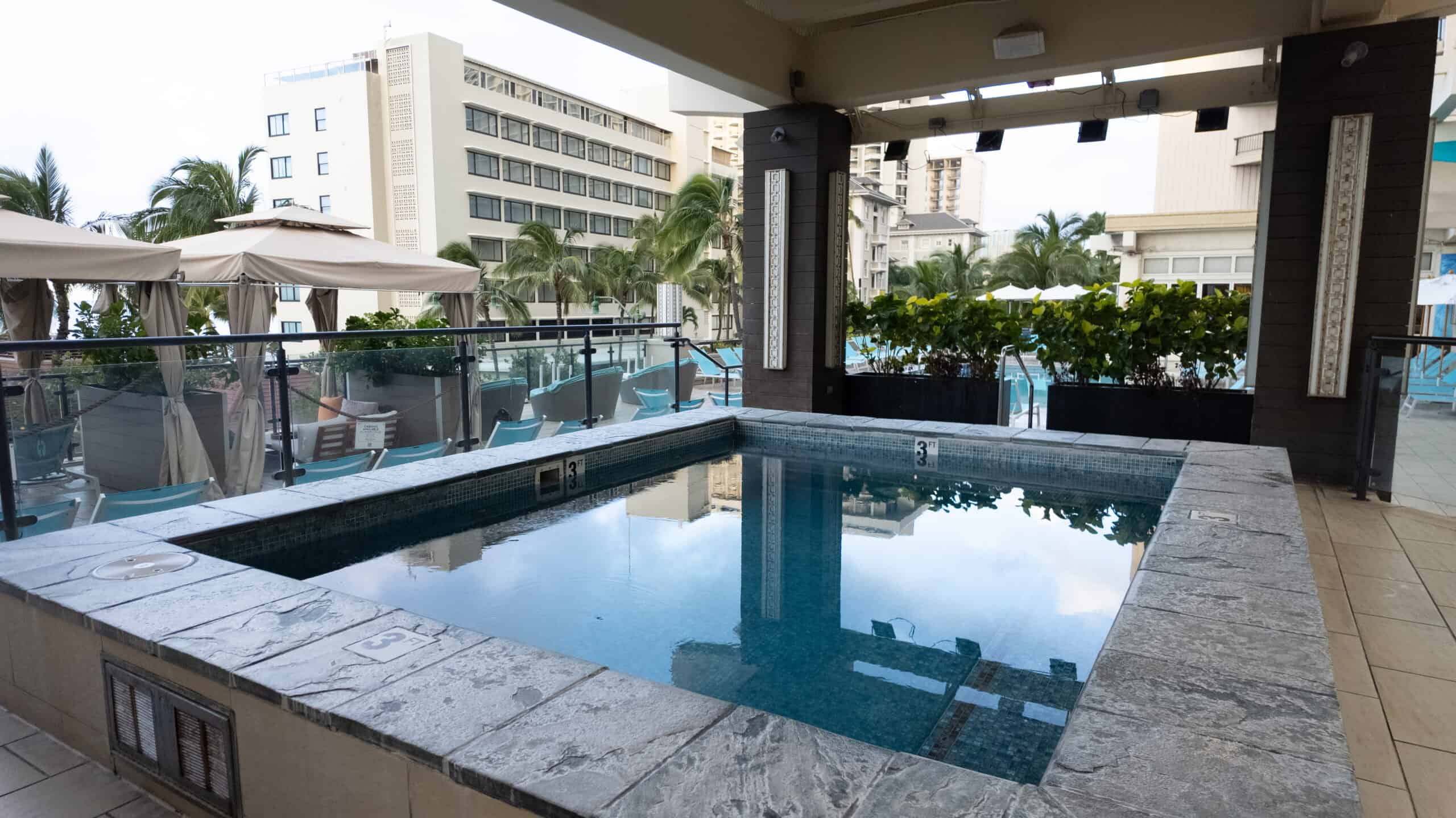 A hot tub with tropical plants, white umbrellas and a pool in the background