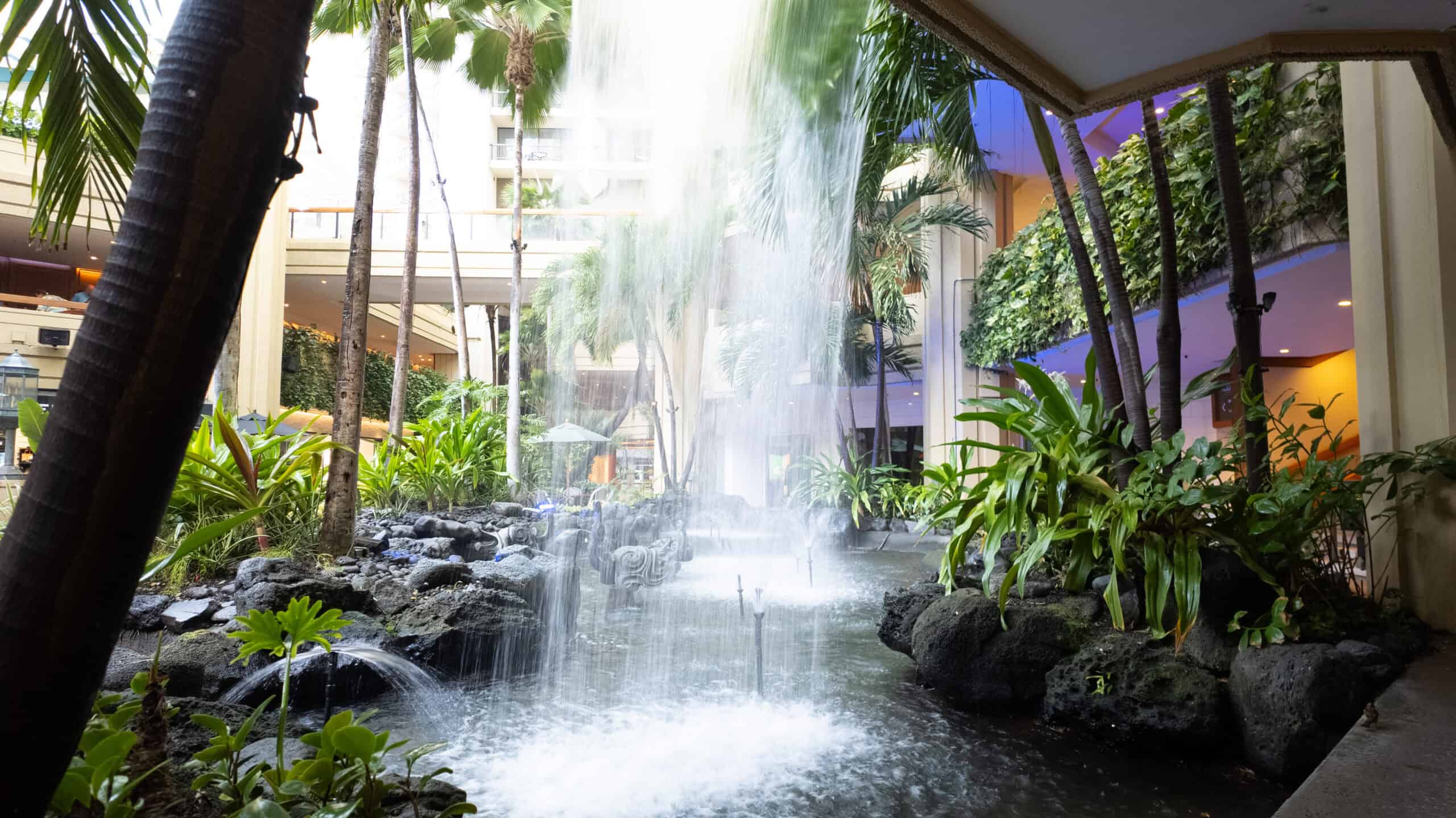 Looking out from behind a waterfall towards tropical plants and an indoor atrium