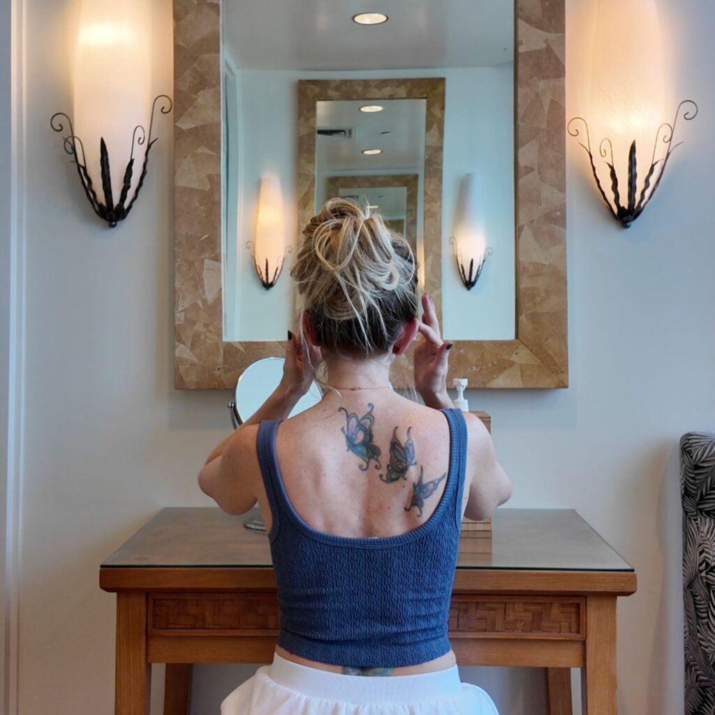 A blonde woman in a blue top looking into a mirror fixing her makeup.