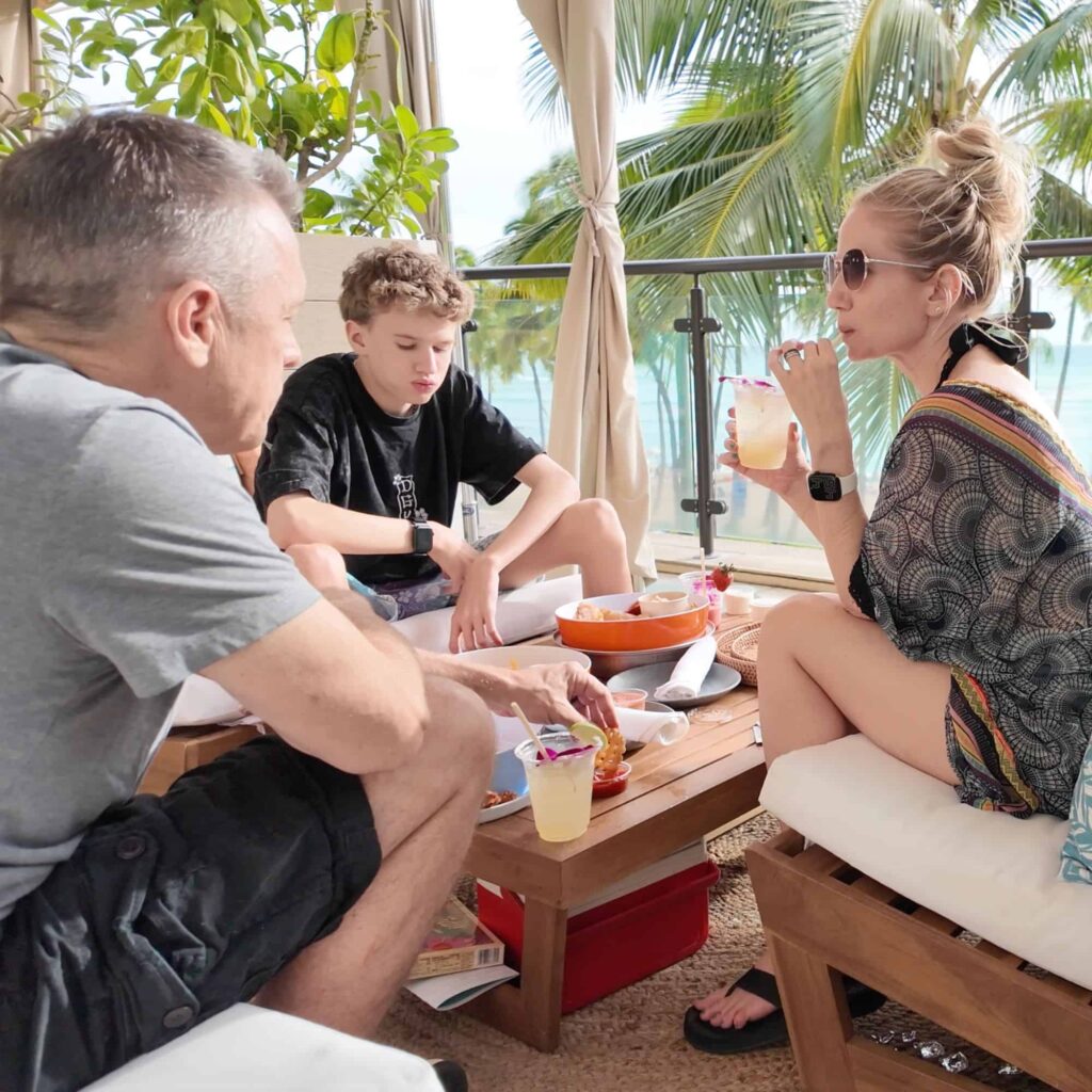 A family sitting at a table with food and drinks, and a beach with palm trees in the background.