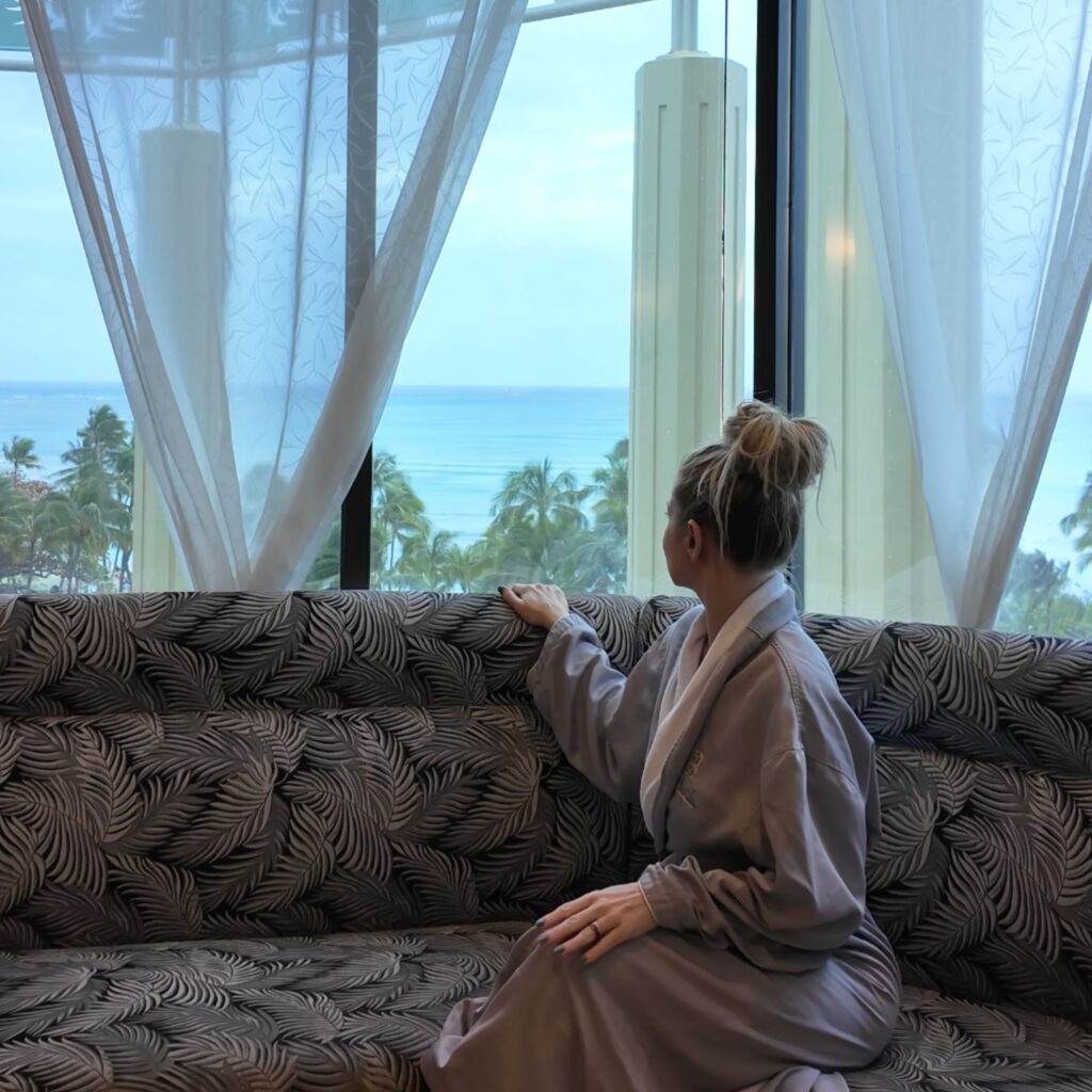 A woman in a gray robe sitting on a bench looking out a window towards the ocean.