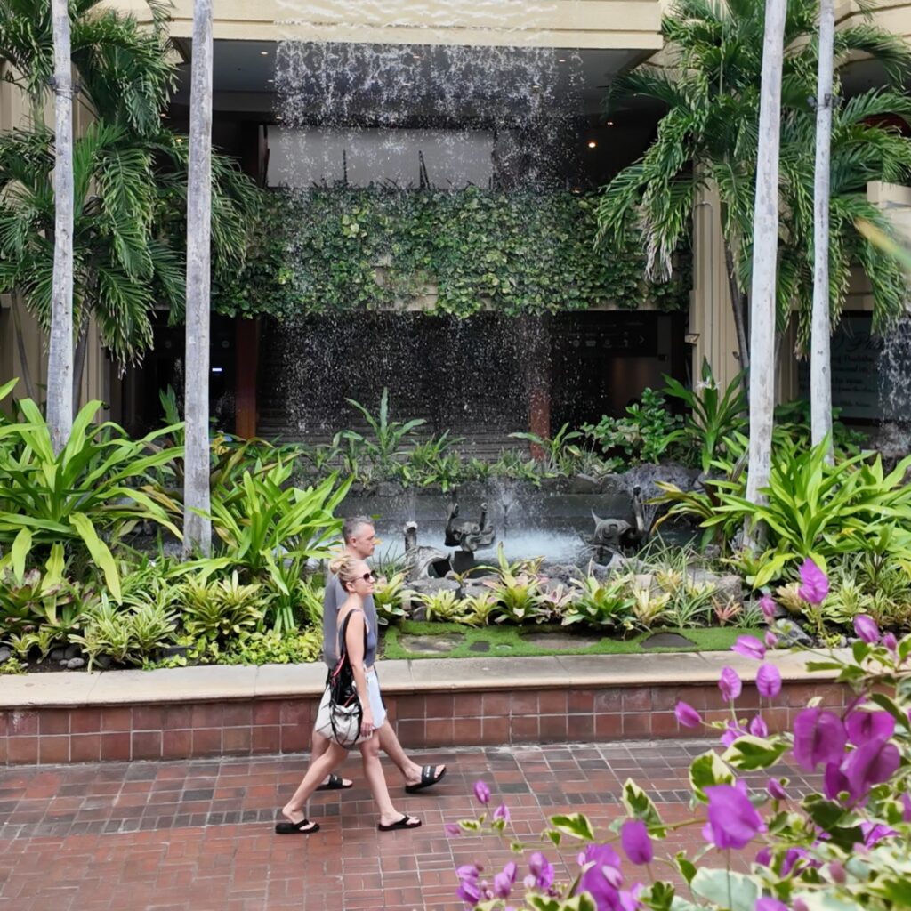 A man and woman walking in front of a waterfall with tropical plants and flowers all around them