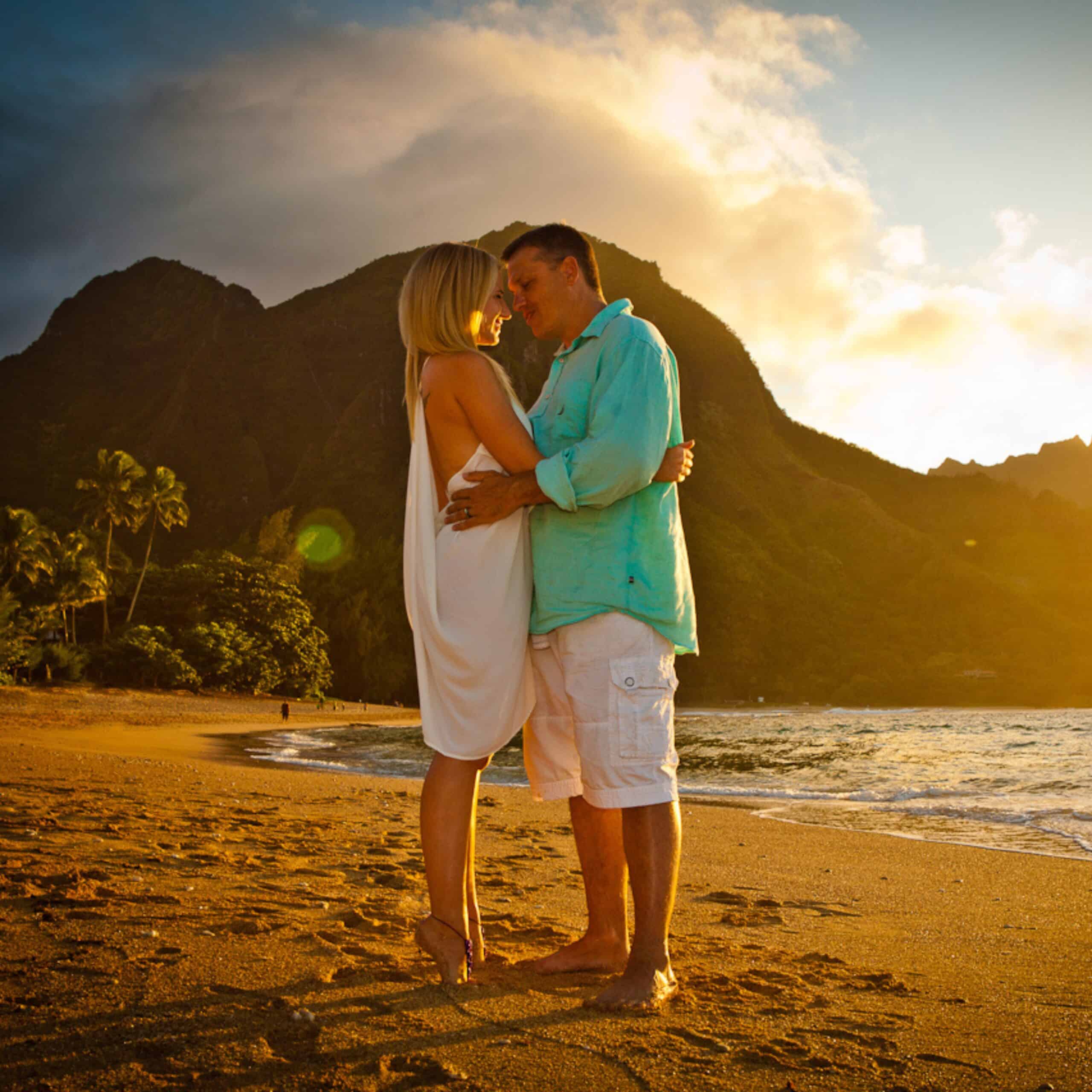A blonde woman in a white wedding dress and man in a turquoise shirt embracing on Tunnels Beach in Kauai