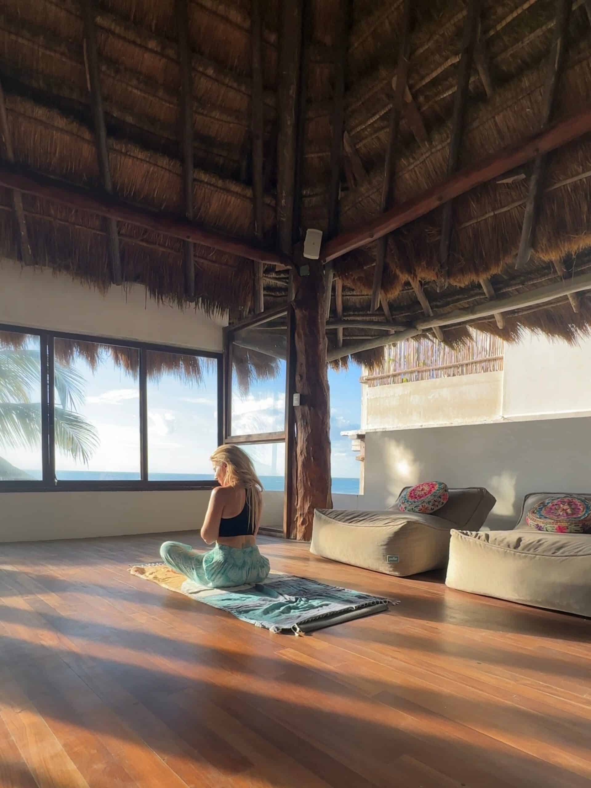 A blonde woman seated on a yoga mat in a tropical studio with palm trees and the ocean out the window in front of her.
