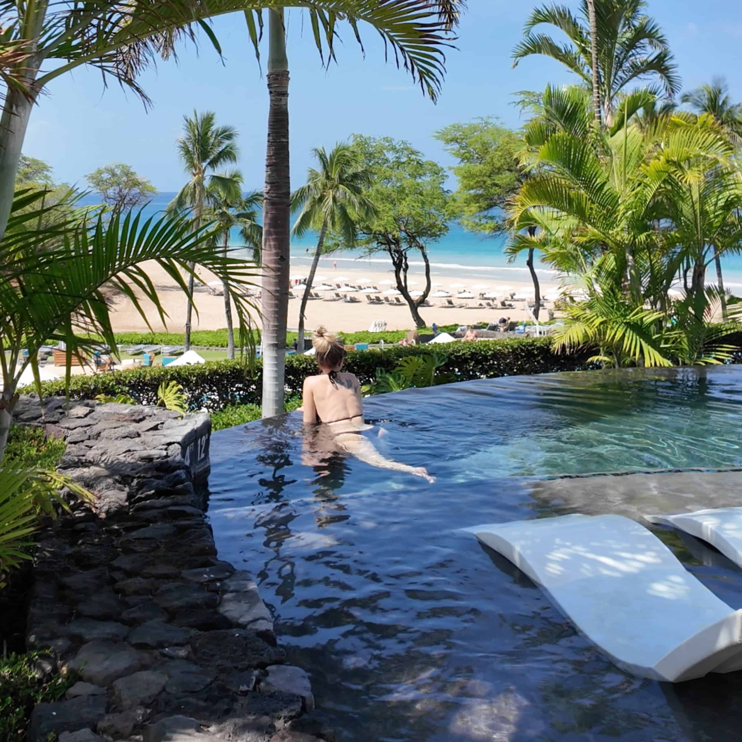 A blonde woman in an infinity pool overlooking a beach in Hawaii surrounded by palm trees