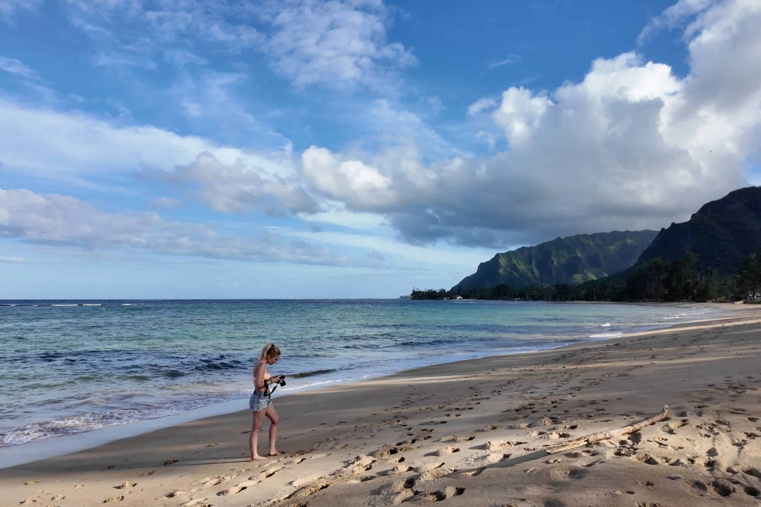 A blonde woman in shorts and a bathing suit on a beach in Oahu looking down at her camera