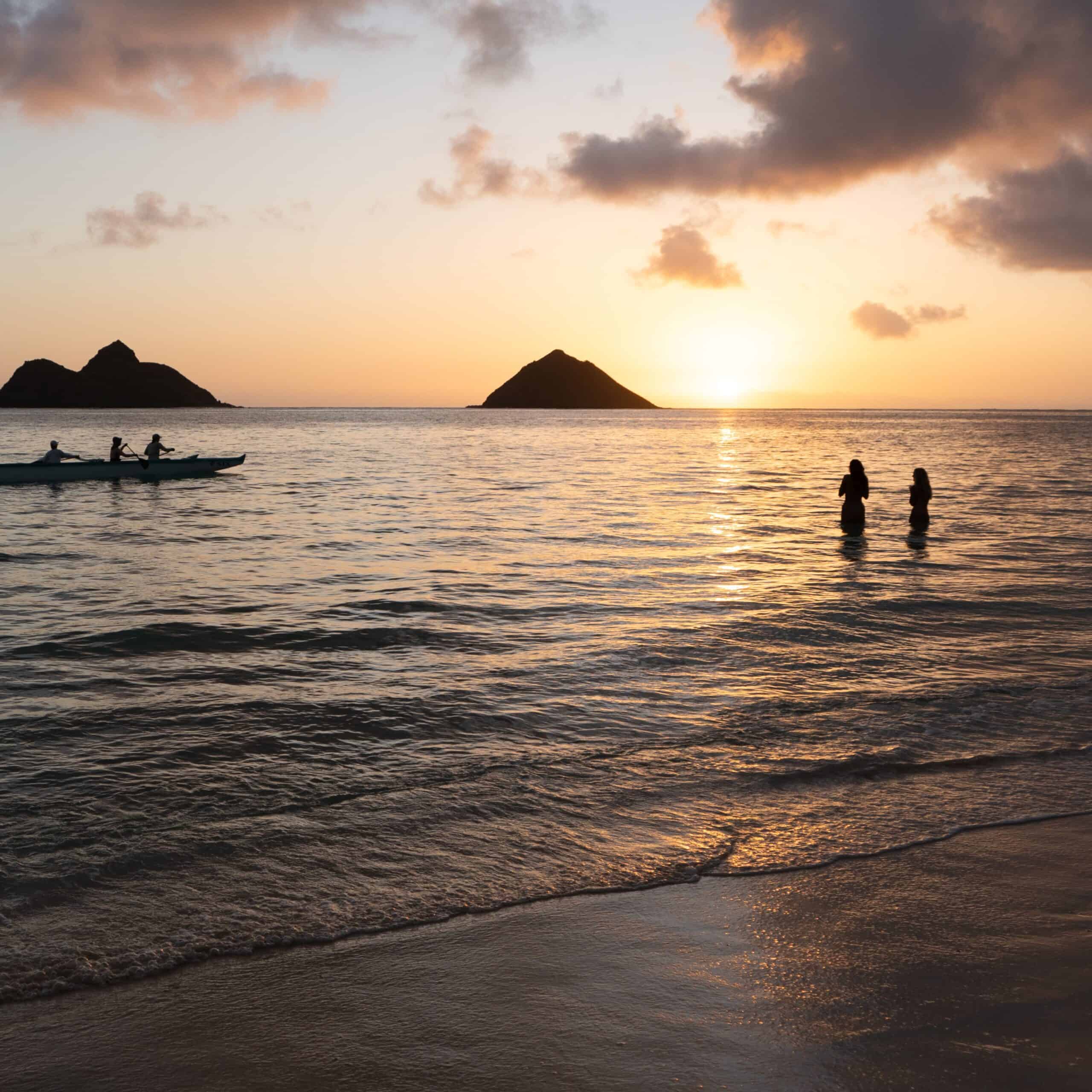 The sun rising over the mokes at Lanikai beach in Oahu with a canoe and people standing in the ocean