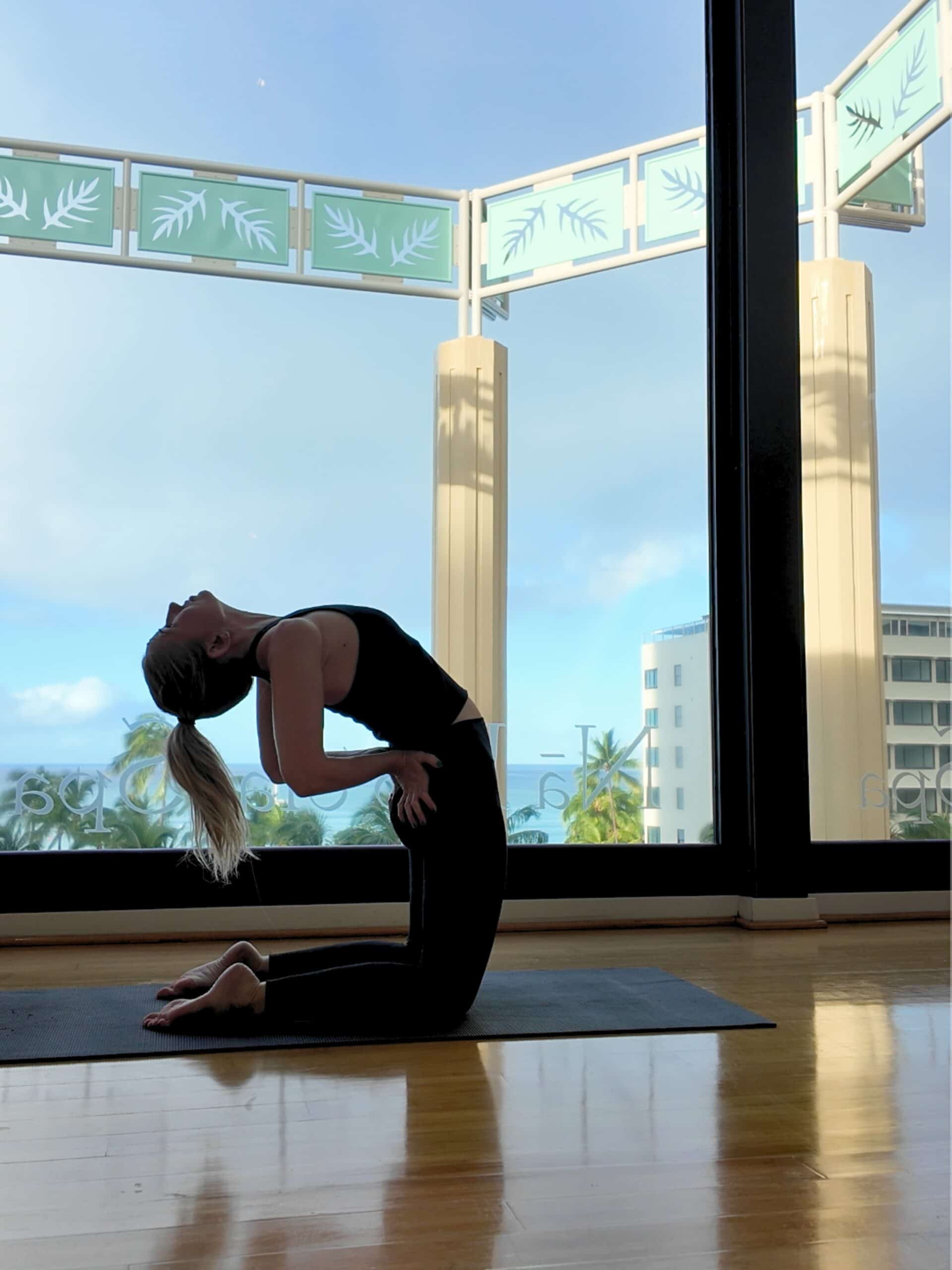 A blonde woman doing a yoga backbend in from of a window looking onto Waikiki Beach