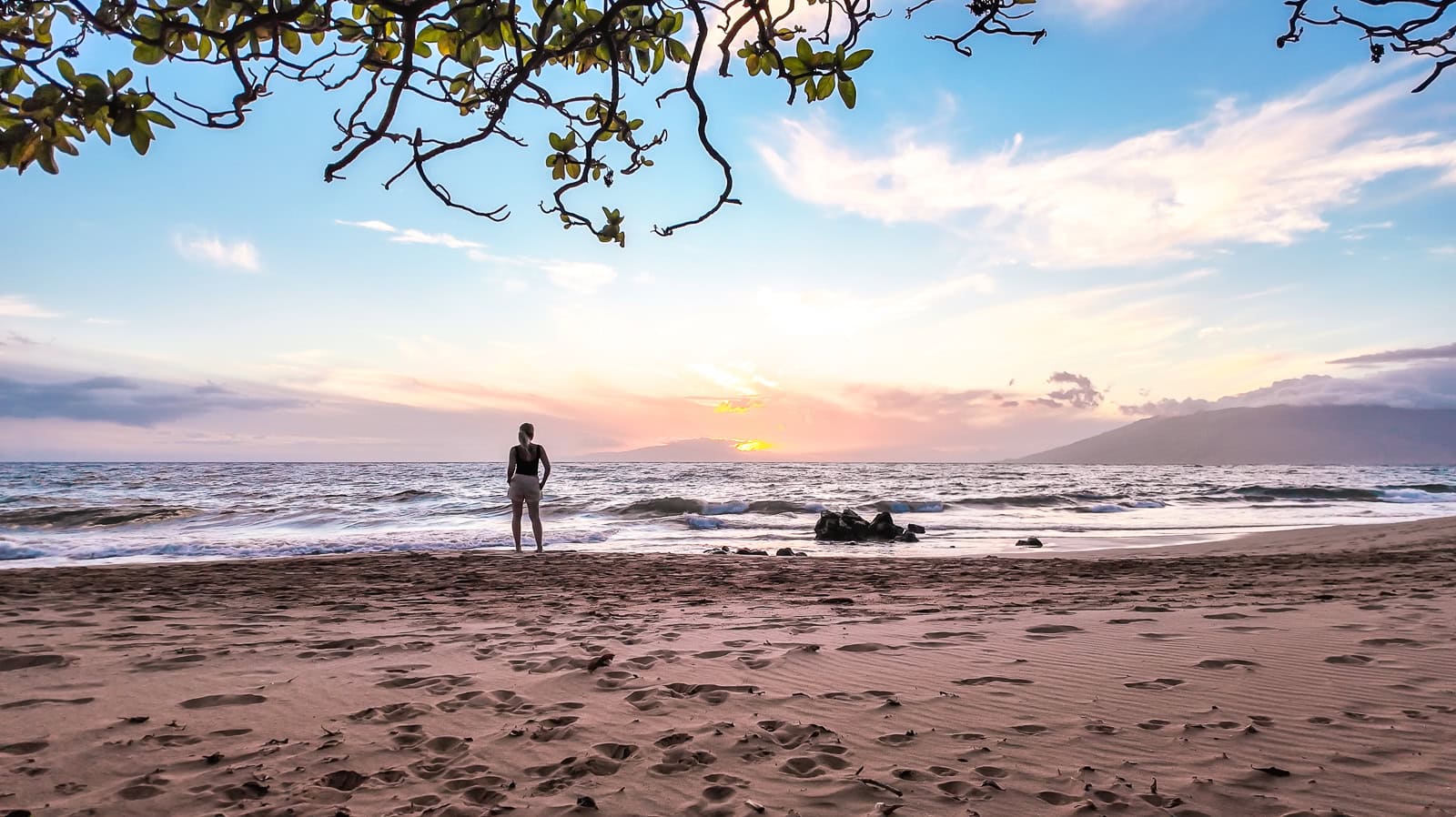The sun setting over the ocean at Keawakapu Beach with waves crashing on the shore and the shadow of a woman standing at the waters edge